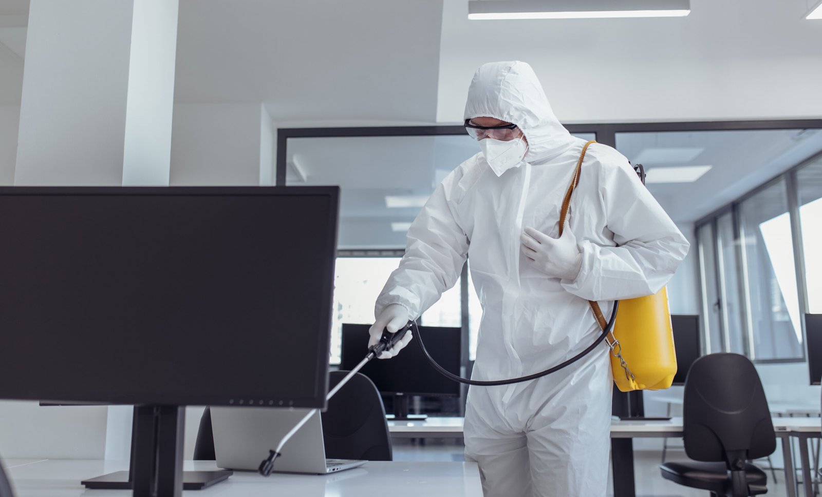 Office disinfection during COVID-19 pandemic. Man in protective suit and face mask spraying for disinfection in the office